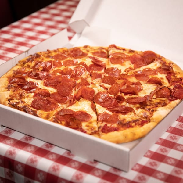 A large, hot pepperoni pizza in an open white box on a red and white checkered tablecloth.